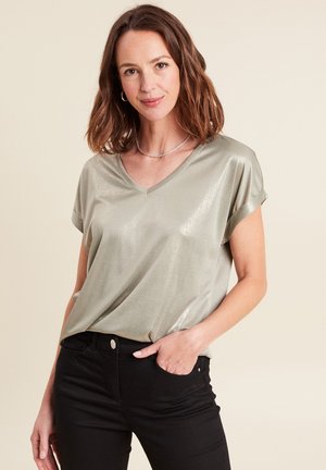 Frau mit schulterlangem braunem Haar trägt ein glänzendes hellgraues, kurzärmeliges V-Ausschnitt-Oberteil und schwarze Hose, steht mit einer Hand in der Tasche.