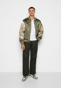 Veste bomber matelassée en vert olive avec texture matelassée, accents argentés, poignets et ourlet côtelés. Associée à un pantalon sombre et des baskets.