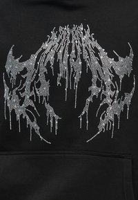 Αφηρημένη σχεδίαση κρανίου από μικρές ασημένιες στρας σε μαύρο ύφασμα, πάνω από μια ραμμένη τσέπη.