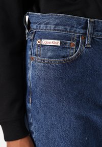 Jeans de mezclilla azul con una superficie texturizada, que presentan una etiqueta de la marca Calvin Klein y remaches de cobre, costuras visibles y un ajuste de media altura.