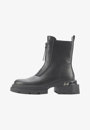 Černá kožená kotníková bota s plochou platformou, bočním zipem a kovovým akcentem poblíž paty. Hladká textura a minimalistický design.