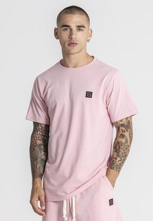 Homme aux cheveux courts et tatouages sur les deux bras, portant un T-shirt rose clair et un short assorti avec des logos carrés noirs.