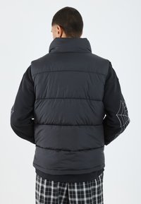 Gilet matelassé noir avec col haut, design rembourré et texture douce. Porté par-dessus une chemise à manches longues noire avec un motif de toile d'araignée.