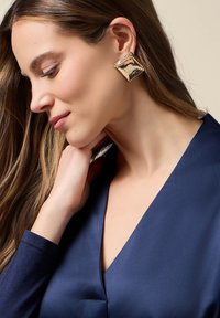 Mujer con cabello largo y castaño, vistiendo un top de seda azul oscuro y grandes pendientes cuadrados dorados, inclinando la cabeza con la mano cerca del cuello.