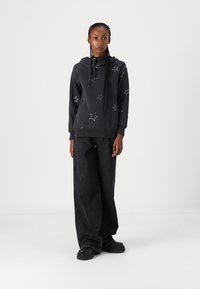 Sudadera negra con patrones de estrellas plateadas, capucha con cordón y bolsillo tipo canguro. Combinada con jeans anchos negros y zapatos negros gruesos.