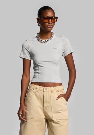 T-shirt grigia aderente con maniche corte, dal tessuto morbido, abbinata a pantaloni a vita alta beige a gamba larga e occhiali da sole marroni oversize.
