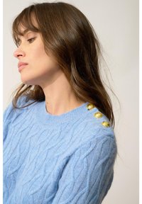 Jasnoniebieski sweter z dzianiny w splot warkoczowy z złotymi dekoracyjnymi guzikami na ramieniu, z okrągłym dekoltem i miękkim, fakturowanym materiałem.