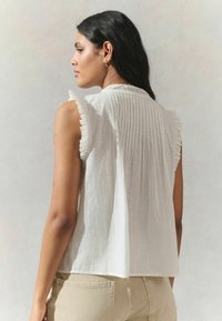 Sleeveless white blouse with ruffled edges and vertical pleats at the back; lightweight, textured fabric, featuring a lace detail near the neckline.