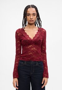 Top de renda burgundy de mangas longas com um decote em V escallopado, padrões florais transparentes e design justo, combinado com jeans escuros.