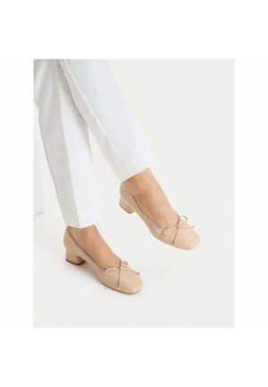 Beżowe baleriny na niskim obcasie z kokardką, noszone z białymi spodniami o prostych nogawkach, na jednolitym jasnym tle.