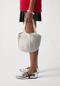 Borsa bianca intrecciata con forma strutturata e due manici. Il materiale è texturizzato, messo in risalto da un maglione rosso a contrasto e scarpe metalliche.