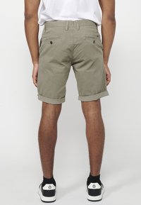 Man in opgerolde olijfgroene shorts, zwart-witte sneakers en een wit t-shirt, staand tegen een witte achtergrond.