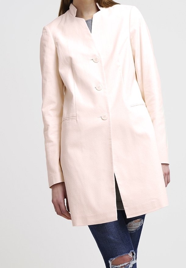 Abrigo entallado rosa claro con cuello mandarín, que presenta cuatro botones, bolsillos laterales y un corte recto, combinado con jeans oscuros rasgados.