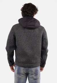 Giacca grigia a spina di pesce con cappuccio isolato nero, caratterizzata da una vestibilità rilassata e tessuto testurizzato. Jeans in denim casual visibili sotto.