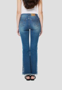 Kék, szárában bővülő farmer, rojtos szegéllyel, oldalán részletes varrással és egy bőr címkével a hátsó részen. Fekete szandállal viselve.