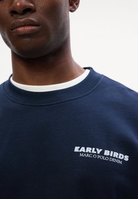 Granatowa bluza z ribbowym białym dekoltem okrągłym. Posiada białą nadrukowaną grafikę "EARLY BIRDS MARC O POLO DENIM" na piersi. Miękka faktura materiału.