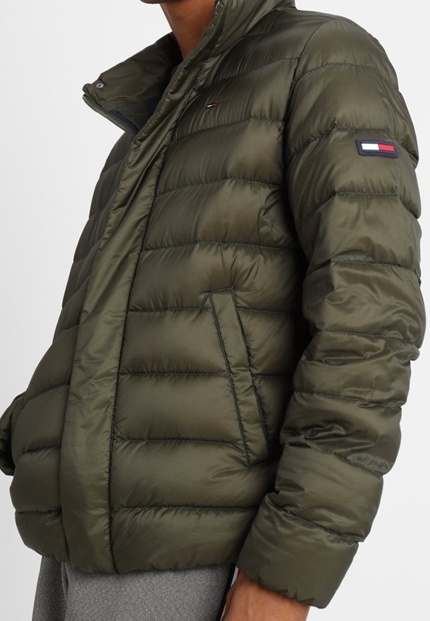 Vadderad olivgrön jacka med hög krage, dragkedjeslutning, dragkedjefickor på sidorna och en logotyp-patch på vänster ärm. Texturerad, kviltad design.