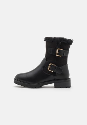 Black ankle boot made of leather and suede, featuring a faux fur trim, gold buckles, and a textured sole.