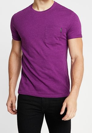 Homme portant un t-shirt violet ajusté à manches courtes avec une poche poitrine et un pantalon noir, debout devant un fond uni.