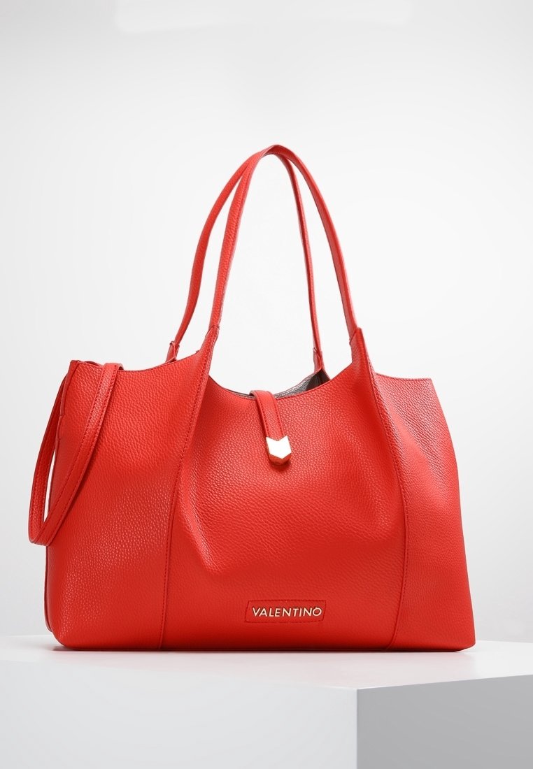 Punainen nahkainen käsilaukku, jossa on teksturoitu pinta, kaksoiskahvat, vetoketjukiinnitys ja kultasävyinen logoleima edessä.