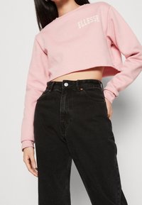 Felpa corta rosa con logo "ELLESSE", abbinata a jeans neri a vita alta. Texture morbida e design a vestibilità rilassata.