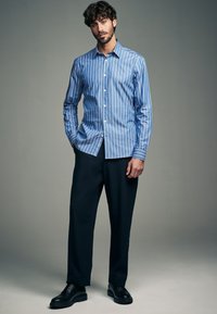 Hellblaues Hemd mit weißen vertikalen Streifen, kombiniert mit dunkler Hose und schwarzen Schuhen, vor neutralem Hintergrund präsentiert.