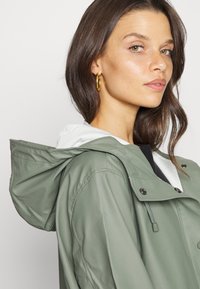 Grüne wasserdichte Jacke mit einem weißen Kapuzenfutter, Reißverschluss und Druckknopfverschlüssen, verstellbarem Kordelzug und auffälligen Nähdetails.
