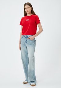 Röd kortärmad t-shirt med vit text i mitten, kombinerad med ljusblå vida jeans och ljusa skor.