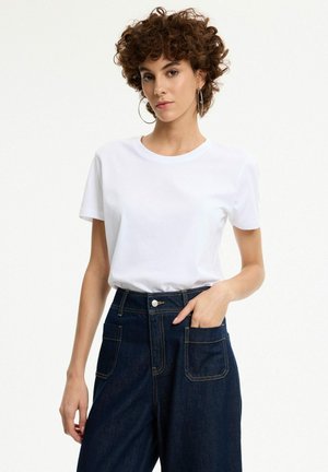 Mujer con cabello corto y rizado que lleva pendientes de aro, una camiseta blanca lisa y vaqueros azul oscuro de cintura alta con bolsillos delanteros.
