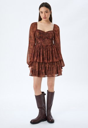 Joven mujer con un vestido marrón de encaje largo y mangas transparentes, con falda en capas, combinado con botas de cuero marrón oscuro hasta la rodilla, de pie sobre un fondo blanco.