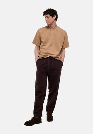 Homme portant un t-shirt beige à manches courtes, un pantalon marron foncé avec cordon et des chaussures marron foncé, debout les mains dans les poches, regardant à gauche.