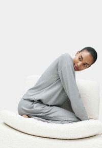 Frau in grauem geripptem Loungewear, die seitlich auf einem strukturierten weißen Sessel sitzt und mit neutralem Ausdruck über die Schulter blickt.