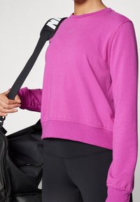 Sudadera morada con cuello redondo, mangas largas y dobladillo acanalado. Presenta un sutil logo de Nike en color gris. Se ve una correa de bolsa negra.