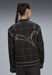 Musta farkkutakki valkoisella ruutukuviolla, jossa on suuri valkoinen Puma-logo selässä, kontrastiväriset ompeleet ja terävä kauluksen muotoilu.