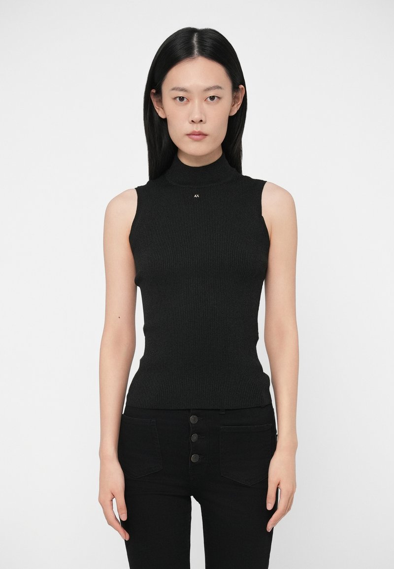 Egyenes fekete hajú nő, aki ujjatlan fekete garbót és magas derekú fekete nadrágot visel, amelyen három gomb található.