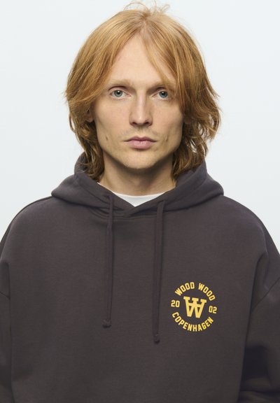 Jeune personne avec des cheveux roux mi-longs portant un sweat à capuche sombre avec le logo "Wood Wood Copenhagen 2002" sur la poitrine, faisant face à l'avant sur un fond uni.