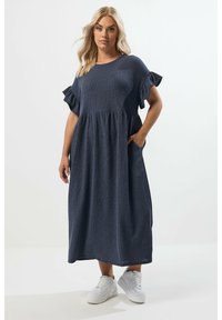 Dunkelblaues, geripptes Maxikleid mit kurzen Rüschensäumen, rundem Ausschnitt, seitlichen Taschen und einem ausgestellten Rock. Kombiniert mit weißen Sneakers.