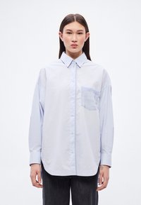 Svetlomodrá, nohavicová košeľa vo veľkom prevedení s golierom a jedným náprsním vreckom. Hladká látka so slabým leskom, zaoblený lem a dlhé rukávy.