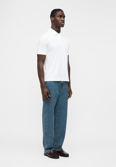 Homme debout portant un polo blanc à manches courtes, un jean bleu et des chaussures habillées marron foncé, sur un fond clair uni.