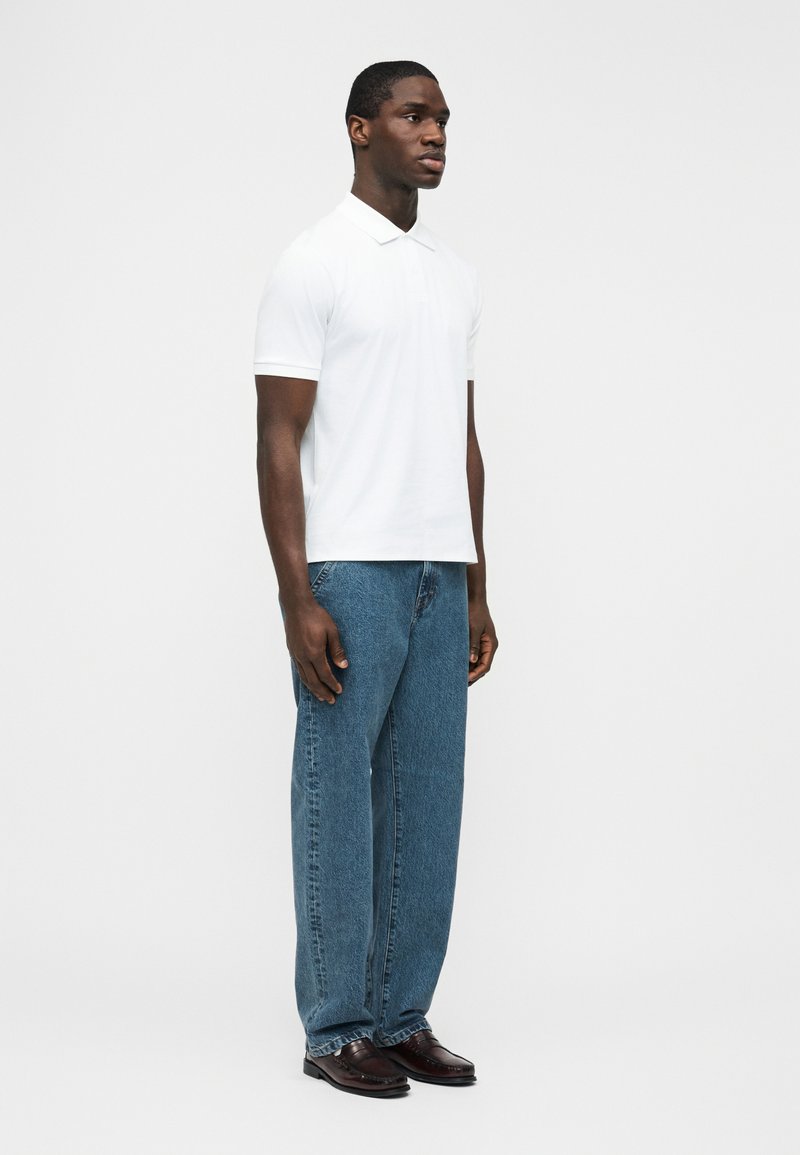 Man standing wearing a white short-sleeve polo shirt, blue jeans, and dark brown dress shoes against a plain light background.