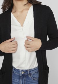 Mustaknit cardigan mustat pitkähihaisilla hihoilla ja sivutaskuilla, kerrostettuna valkoisen kauluksettoman paidan päälle. Cardiganissa on pehmeä pinta ja auki oleva etuosa.