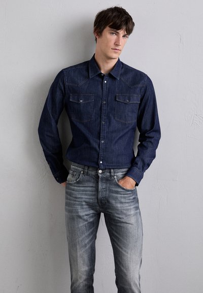 Camicia in denim blu scuro con yoke a punta, due tasche sul petto e colletto button-down. Abbinata a jeans grigi chiari sbiaditi.