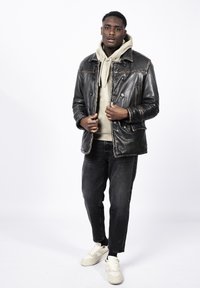 Chaqueta de cuero negra con detalles de botones, sudadera con capucha verde oliva debajo, jeans de mezclilla oscura y zapatillas de color claro.
