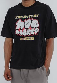 Black cotton t-shirt with oversized graphic text in white, red, and yellow. Short sleeves and a relaxed fit, featuring a round neckline.