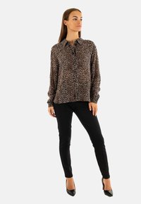 Blusa de estampado de leopardo con mangas largas y cierre de botones, combinada con pantalones ajustados negros y tacones altos negros. Tejido suave con brillo.