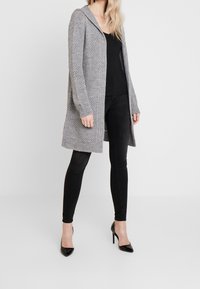 Grå stickad cardigan med huva, öppen front, ribbade ärmslut och fåll. Bärs över en svart topp och kombineras med svarta slim-fit jeans och klackar.