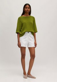 Groene gebreide trui met wijde mouwen, gecombineerd met witte op maat gemaakte shorts. Het model draagt doorzichtige ballerina's en staat tegen een effen achtergrond.