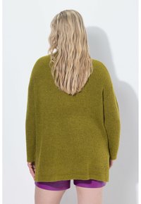 Maglione di maglia oversize verde oliva con tessuto testurizzato, spalle scese e orlo curvo. Il modello indossa pantaloni corti viola sotto.