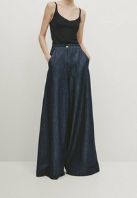 Femme portant un haut sans manches noir et un pantalon en denim large de couleur foncée, les mains dans les poches, se tenant contre un fond uni.