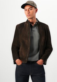 Brown suede jacket with a high collar, silver zipper, worn over a grey polo shirt, paired with dark denim jeans. The cap features a checked pattern.
